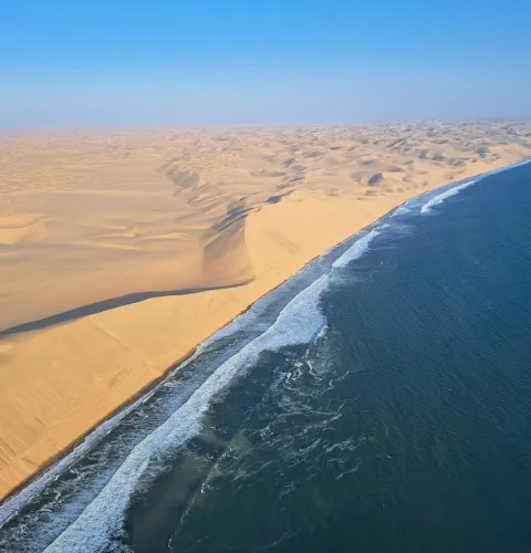 Aerial view from a plane of the South Atlantic Ocean meeting the Namib Desert on an unpopulated stretch of coastline, dune mounts visible in the distance