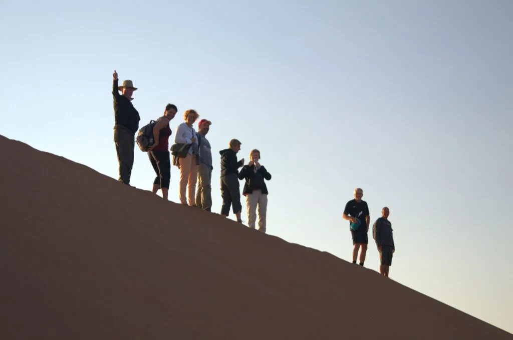 Group of tourists silhouetted on a sand dune at sunset, one waving at the camera